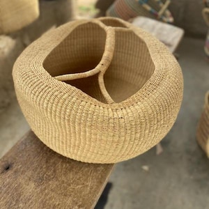 May include: A large, round woven basket with three interior compartments and a handle. The basket is made of natural-colored fibers and sits on a wooden surface. The basket's design is simple and functional.