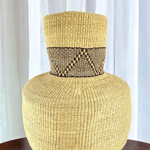 May include: A large, round, woven basket with a brown and tan geometric pattern around the top. The basket is made of natural fibers and has a light tan color.
