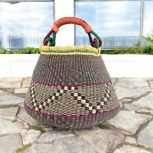 May include: A handwoven basket with a rounded shape and a sturdy handle. The basket features a geometric pattern in black, green, and purple, with a tan border. The handle is wrapped in brown leather and green and purple rope.