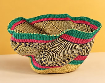 May include: A woven basket featuring a flexible, organic shape with a wide, undulating rim. The basket is constructed from natural tan fibers accented with bold, horizontal bands of vibrant green and hot pink, plus dark geometric patterns.