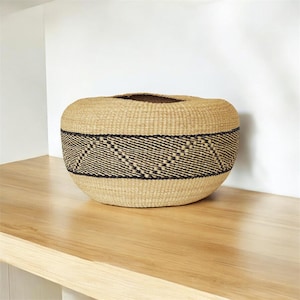 May include: A large, round, woven basket with a black and brown geometric pattern around the middle. The basket is made of natural fibers and has a rustic, handmade look.