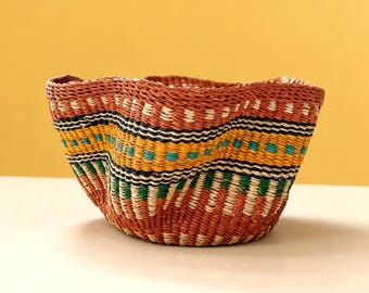 May include: A round, woven basket featuring a wavy, flexible rim. The exterior displays a repeating pattern of horizontal bands in terracotta, yellow, black, white, and turquoise green, created with tightly coiled, natural fiber strands.