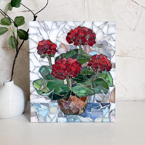 May include: A rectangular mosaic art piece featuring a potted plant with vibrant red flower clusters and dark green leaves. The pot is rendered in shades of brown, set against a background of white and light blue glass tiles with visible grout lines.