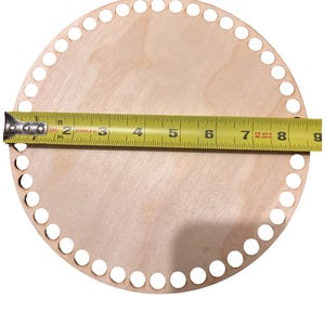 May include: A round, unfinished wooden base with evenly spaced holes around the perimeter. A yellow measuring tape is laid across the diameter, showing a measurement of approximately 8 inches. The base is likely for a basket or craft project.