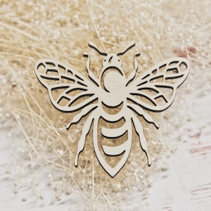 May include: A wooden bee-shaped cutout with intricate wing and body details. The bee is light-colored, possibly unfinished wood, and rests on a bed of dried, beige-colored flowers and stems. The background is a light-colored wooden surface.