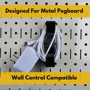 Puede incluir: Organizador negro para panel perforado que sostiene un adaptador de corriente y un cable blancos. Diseñado para paneles perforados metálicos y compatible con Wall Control. Texto "Designed For Metal Pegboard" y "Wall Control Compatible" en barras amarillas.