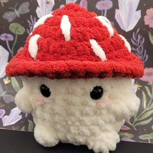 May include: A plush mushroom toy with a red cap featuring white accents. The mushroom has a white body, black eyes, and pink cheeks. The background is a dark pattern with mushrooms and crystals.