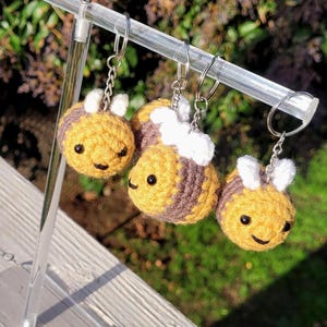 May include: Three handmade crochet bee keychains. Each bee features a yellow body with brown stripes, white wings, and a smiling face. The keychains are attached to silver metal rings and are displayed on a clear acrylic stand.