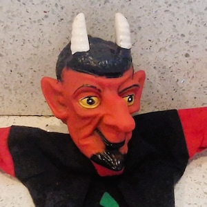 May include: A devil hand puppet with a red face, black beard, and white horns. The puppet has a black body with red hands and green accents. The puppet's face has yellow eyes and a sinister grin.