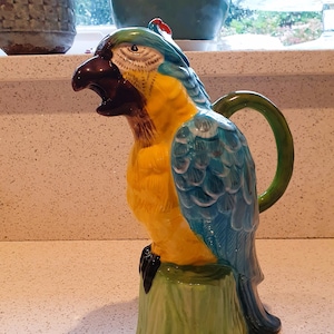 May include: A ceramic parrot pitcher with a blue and yellow body, a green perch, and a green handle. The parrot has a black beak and a red crest.