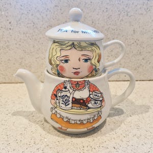 以下が含まれることがあります： 女の子が紅茶とケーキを運んでいる漫画のイラストが描かれた白い陶器製のティーポットとティーカップのセット。ティーカップには蓋があり、蓋には「TEA for MOM」の文字が書かれています。