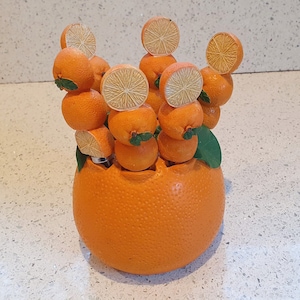 Puede incluir: Soportes de cerámica en forma de naranja con hojas verdes, dispuestos en un cuenco de cerámica más grande en forma de naranja. Los soportes están diseñados para sostener palillos de dientes o palitos de cóctel.