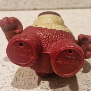 Peut inclure: Figurine jouet rouge et beige avec une surface textur&eacute;e. La figurine a deux pieds rouges avec des indentations circulaires et du texte. La figurine a deux bras et une t&ecirc;te avec un dessus beige.