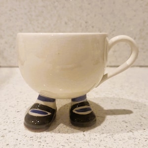 Puede incluir: Una taza de cerámica de color crema con asa, apoyada sobre dos pies. Los pies están pintados con zapatos negros, detalles azules y calcetines a rayas azules y blancas. La taza tiene forma redondeada.