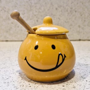 Könnte beinhalten: Ein gelber Keramik-Honigtopf mit einem Smiley-Gesicht. Der Topf hat einen Deckel und einen kleinen Honiglöffel aus Holz. Das Smiley-Gesicht hat schwarze Augen und eine herausgestreckte Zunge. Der Topf steht auf einer gesprenkelten Oberfläche.
