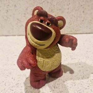 Peut inclure: Figurine d'ours rouge et beige avec une expression triste. L'ours a un grand nez brun fonc&eacute;, un ventre beige et se tient debout, les bras tendus. La figurine est un personnage de la franchise Toy Story.