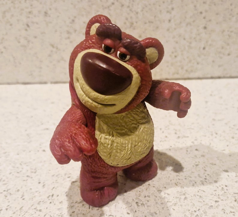 Peut inclure: Figurine d'ours rouge et beige avec une expression triste. L'ours a un grand nez brun fonc&eacute;, un ventre beige et se tient debout, les bras tendus. La figurine est un personnage de la franchise Toy Story.