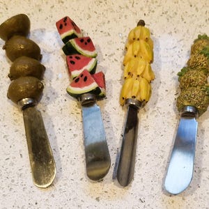 May include: Four decorative cheese spreaders with fruit-shaped handles. The handles feature kiwi, watermelon, bananas, and pineapple designs. The blades are silver-colored and the fruit is painted in realistic colors.