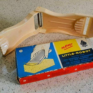 以下が含まれることがあります： ハンドルとブレードが付いた白いプラスチック製のキッチンツール。ツールには「Little-Robot」というラベルが貼られており、「すべての主婦のためのユニバーサルキッチンツール」と説明されています。