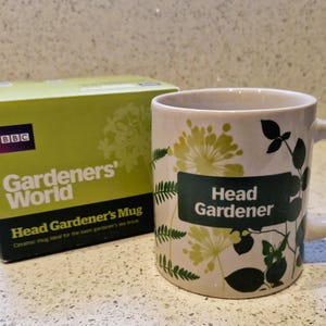 Puede incluir: Taza de cerámica con la inscripción "Head Gardener" en una etiqueta verde, rodeada de diseños florales y hojas. Junto a ella, una caja verde con el logotipo de "Gardeners' World".