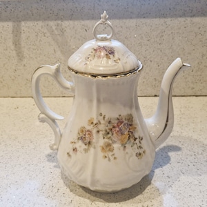 May include: White porcelain coffee pot with a lid, handle, and spout. The pot is decorated with floral patterns in shades of yellow, pink, and purple. Gold trim accents the rim and handle. The lid has a small decorative finial.