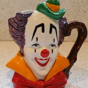 Puede incluir: Una taza de cerámica de payaso con un sombrero verde, pelo morado, una pajarita roja y un asa marrón.