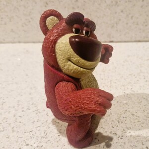 Peut inclure: Un ours en peluche rouge avec un museau beige et un nez marron fonc&eacute;. L'ours a une expression amicale et est pos&eacute; avec les bras tendus. Le jouet est un personnage d'un film d'animation populaire.