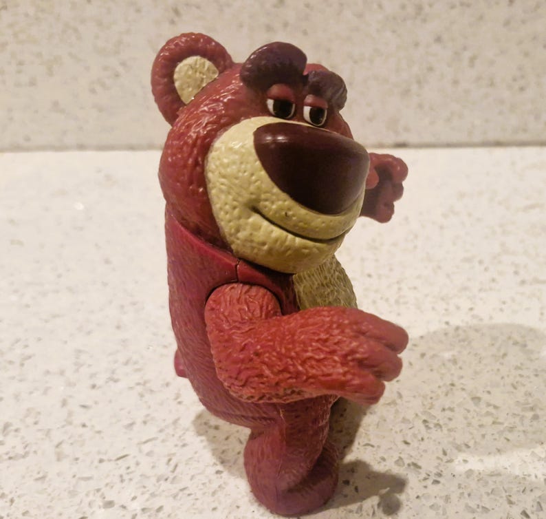 Peut inclure: Un ours en peluche rouge avec un museau beige et un nez marron fonc&eacute;. L'ours a une expression amicale et est pos&eacute; avec les bras tendus. Le jouet est un personnage d'un film d'animation populaire.