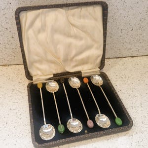 Puede incluir: Un juego de seis cucharas de plata con cuencos facetados y mangos redondeados de colores. Las cucharas se exhiben en un estuche vintage marrón oscuro con interior de terciopelo negro y forro de satén color crema.