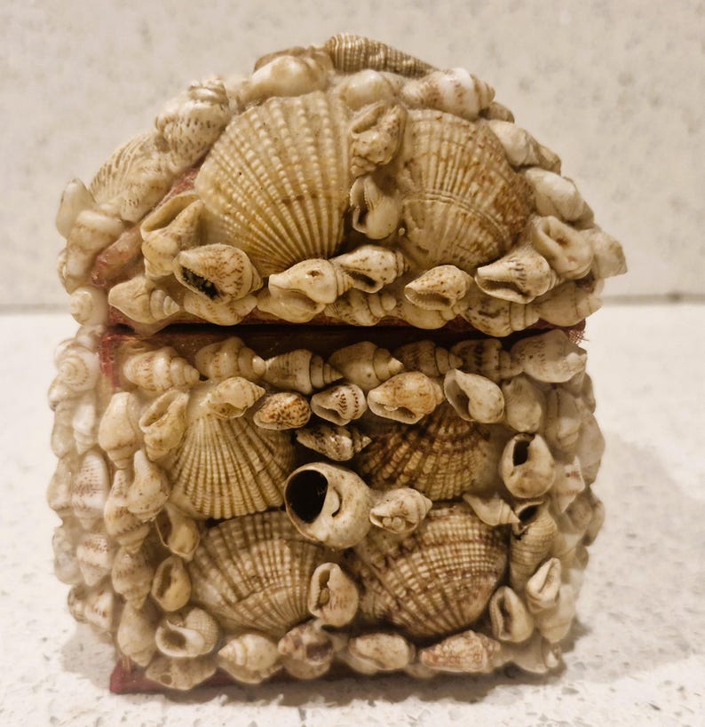 May include: A decorative box covered in various seashells. The box is rectangular with a hinged lid. The seashells are in shades of beige and brown, creating a textured surface. The box is likely used for storing small items.
