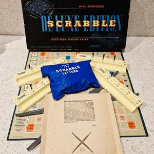 May include: Deluxe Scrabble Edition board game with a black box, beige letter racks, a blue bag for letters, and a game board. The box reads "Deluxe Scrabble Edition" and "With Turntable". The game board has scoring areas.
