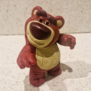Peut inclure: Figurine d'ours rouge et beige avec une expression triste. L'ours a un grand nez brun fonc&eacute;, un ventre beige et se tient debout, les bras tendus. La figurine est un personnage de la franchise Toy Story.