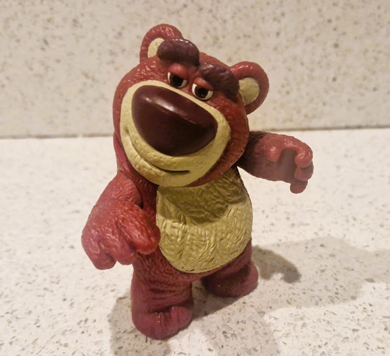 Peut inclure: Figurine d'ours rouge et beige avec une expression triste. L'ours a un grand nez brun fonc&eacute;, un ventre beige et se tient debout, les bras tendus. La figurine est un personnage de la franchise Toy Story.
