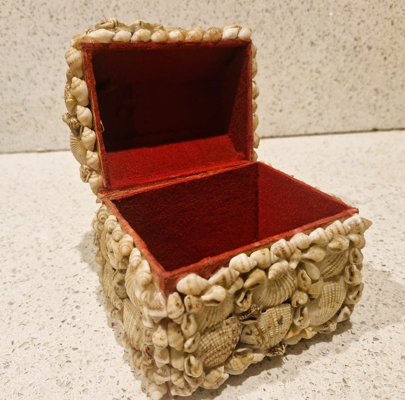 May include: A small jewellery box covered in seashells. The box has a red velvet interior and a hinged lid. The seashells are primarily beige and cream-coloured, creating a textured surface. The box is likely used for storing small items.