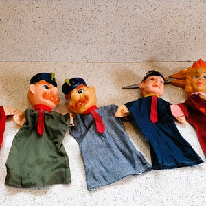 May include: Five vintage hand puppets with painted faces and fabric clothing. The puppets have different outfits in red, green, blue, and gray. One puppet has a yellow collar, and another has a red tie. The puppets are arranged in a row.