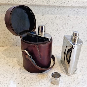 Puede incluir: Una funda de cuero marrón para frasco con cierre de cremallera y correa de transporte. La funda está abierta y contiene un frasco de plata con tapa de rosca. También se muestra un frasco de plata separado y un pequeño vaso de chupito de plata.