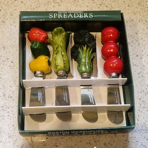 Könnte beinhalten: Ein Set aus vier Brotaufstrichmessern mit Gemüsegriffen, darunter eine rote Paprika, grüne Paprika, gelbe Paprika, Salat, Brokkoli und Tomaten. Die Edelstahlklingen werden in einer grünen Box präsentiert.