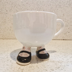Puede incluir: Taza de cerámica blanca con asa y zapatos negros y blancos de lunares para las piernas.