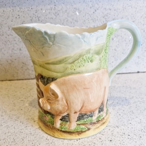 May include: A ceramic pitcher with a detailed relief of a pig. The pitcher features a landscape design with rolling green hills and a light blue sky. The pig is a light pink color and stands on a patch of green grass.