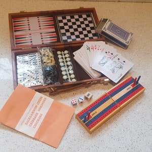 May include: A wooden travel game set in a brown leatherette case. The set includes a checkerboard, dominoes, a cribbage board, playing cards, and dice. The case has the words "Backgammon Chess Cribbage Dominoes Checkers" printed on the front.