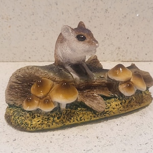 May include: A small, brown and white mouse figurine sitting on a log with brown and white mushrooms. The figurine is made of resin and has a realistic appearance.