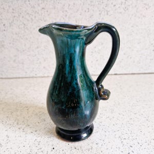 Op de afbeelding: Een kleine keramische kan met een donkergroene en groene glazuur. De kan heeft een gebogen handvat en een tuit. De basis is donker en effen van kleur. De kan heeft een kleine inscriptie op de voorkant.