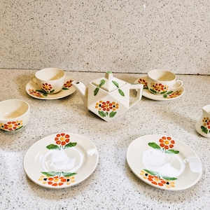 Puede incluir: Un juego de té blanco con diseños florales de color naranja y verde. El juego incluye una tetera, cuatro tazas y una jarra de crema.