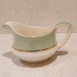 May include: A small, white ceramic creamer with a green band and gold trim. The creamer has a rounded body and a curved handle.