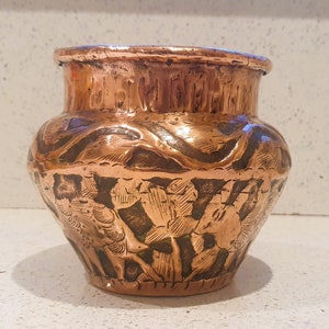 May include: A copper pot with an intricate floral design. The pot is a deep reddish-brown color and has a rounded shape.