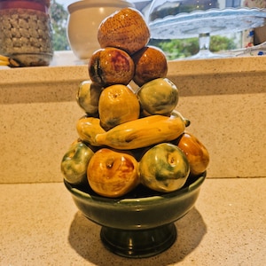Può includere: Una ciotola di frutta in ceramica con una glassa verde e una base a piedistallo. La ciotola è piena di una varietà di frutta in ceramica, tra cui mele, pere, arance e banane.
