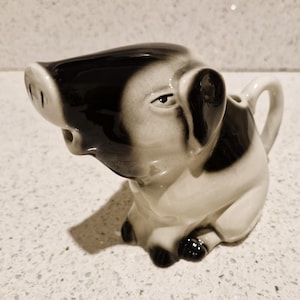 Puede incluir: Un jarro de crema de cerámica con forma de cerdo con esmalte blanco y negro. El cerdo tiene un hocico, ojos y una asa en la parte posterior.