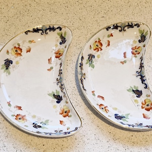 May include: Two white, crescent-shaped decorative plates with a gold rim. Each plate features a floral design with orange, yellow, and blue flowers and green leaves. The plates have a dark blue border with ornate detailing.