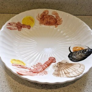 Puede incluir: Plato de servir de cerámica blanca con borde festoneado. El plato está decorado con un cangrejo, dos langostas, una almeja y un mejillón. El plato es perfecto para servir mariscos.