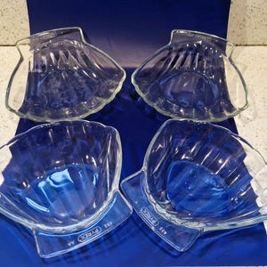 May include: Four clear glass Pyrex shell-shaped bowls. The bowls have a blue tint and a ribbed design. Each bowl has a rectangular base with the Pyrex logo and model number. The bowls are arranged on a blue surface.
